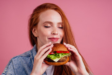 redhaired ginger woman with freckles enjoying big huge burger cutlet and wearing demin american jeans jacket in studio pink background .USA traditional concept