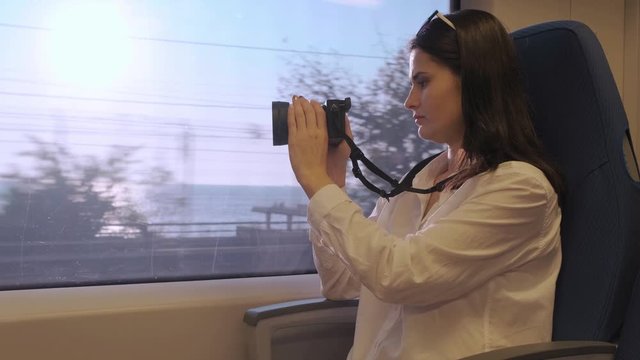 A woman rides a train and takes a photo of the sea from the window on the camera . A tourist girl goes on a Sunny day on a train and takes pictures from the window.