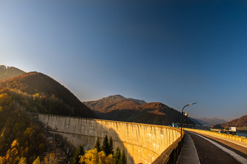 Beautiful view of a dam seen from up high, surrounded by forests and colored trees, in a sunny day in autumn