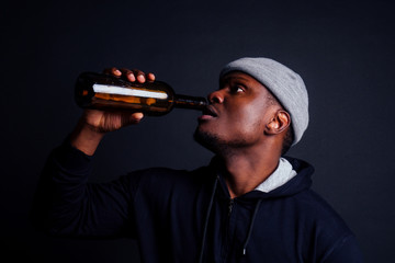 african american gue wearing gray hat and hoody hjlding botle with wine in srudio black background.alcoholism homeless bum concept