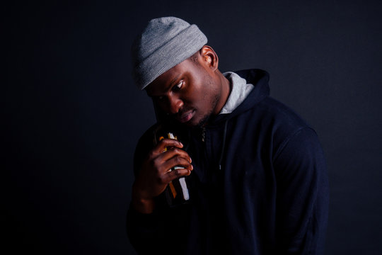african american gue wearing gray hat and hoody hjlding botle with wine in srudio black background.alcoholism homeless bum concept