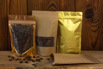 Brown kraft paper and golden metallized pouch bags with coffee beans front view on a wooden background.