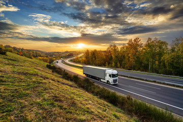 White trucks driving on the asphalt highway in autumn landscape at golden sunset with dramatic clouds
