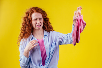 curly ginger redhead woman in a cotton shirt gesture smells bad holding a dirty pink sock out a disgusted look on her face in studio yellow background - obrazy, fototapety, plakaty