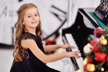 Fototapeta na wymiar little cute girl with a beautiful hairstyle and elegant black dress playing the piano or piano on the background of Christmas decor and a large dial clock.