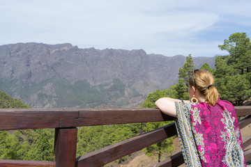 Fototapeta na wymiar Teen tourist resting, hold a wood fence, relaxing and look the environment national park with colorful dress. Blonde woman observe and enjoy the forest landscape in a view point.