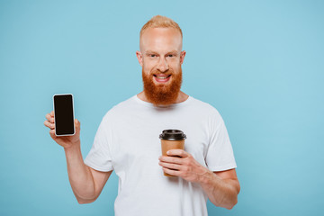 happy bearded man with coffee to go showing smartphone with blank screen, isolated on blue