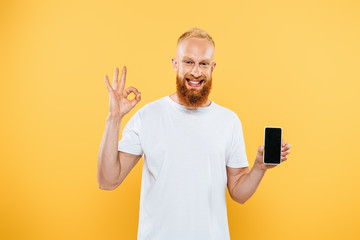 smiling bearded man showing ok sign and smartphone with blank screen, isolated on yellow