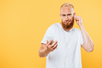 skeptical bearded man listening music with earphones and smartphone, isolated on yellow