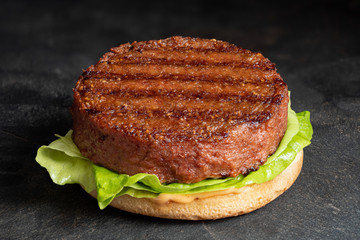 Freshly grilled plant based burger patty on bun with lettuce and sauce isolated on black slate.