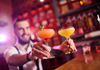 handsome bartender prepares an orange alcoholic cocktail and smiles on the background of a bar or a...