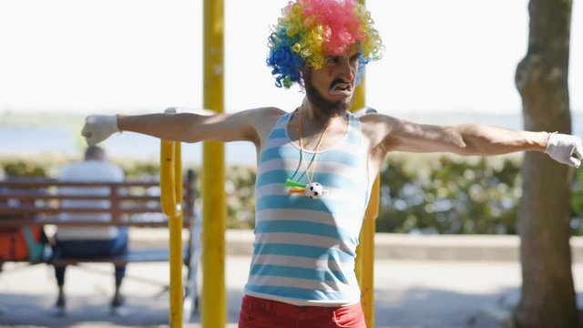 Mime pulls up on the uneven bars in the park. The clown shows the pantomime how he does sports on the street.
