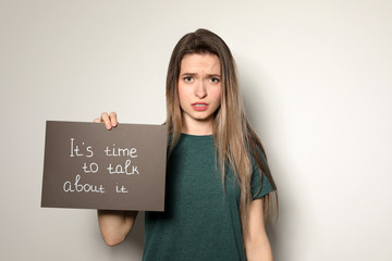 Young woman holding card with words IT'S TIME TO TALK ABOUT IT against light background