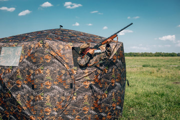 Good shot standing in ambush in the khaki tent, aiming to the sky; steppe and sky; huntsman concept.