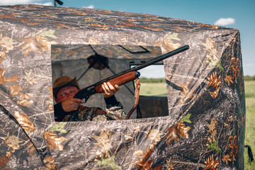 Lucky hunter sitting in wait, the man holds a rifle standing in a camouflage tent wearing a khaki...