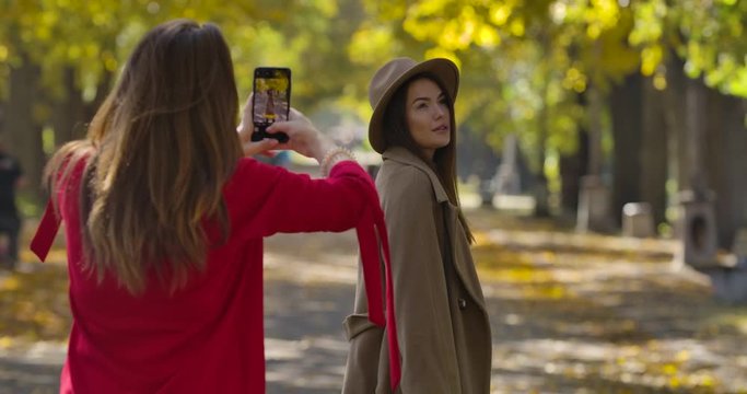 Beautiful Caucasian woman in elegant brown coat and hat posing for her female friend on the background of autumn park. Positive European women taking photos outdoors. Cinema 4k footage ProRes HQ.