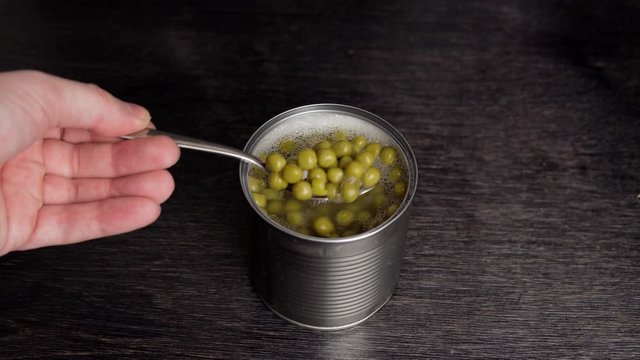 Male hand with small spoon takes canned peas from tin can on black background.