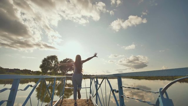 Happy seventeen year old girl in overalls runs with raised hands on a background of sunset and lake.