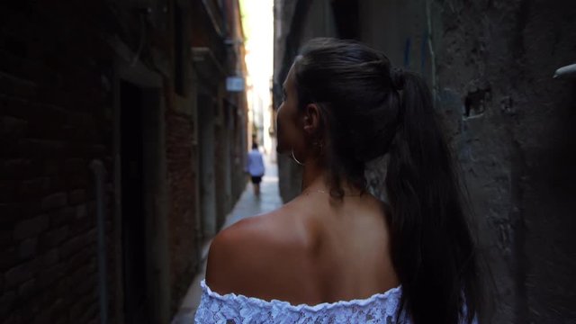 girl with ponytail in summer lacy top walks along narrow shady street in Venice looking at old houses close view