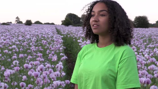 Beautiful happy mixed race African American girl teenager female young woman walking through field of pink poppy flowers taking photographs for social media on her mobile smart phone