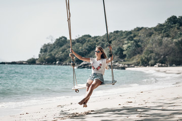 Attractive slim girl on the rope swings, sandy beach against the blue bay, sunny day, relaxation.