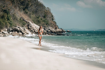 Attractive slim blondie in bikini walks along the sandy beach against the blue bay, sunny day, relaxation.