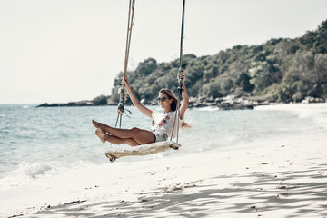 Attractive slim girl on the rope swings, sandy beach against the blue bay, sunny day, relaxation.