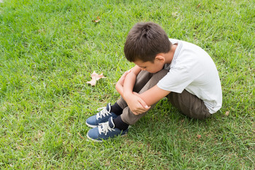 Child sitting on the grass with his chin resting on his knees, loneliness and sadness concepts.