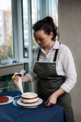 Pastry chef squeezes beige cream on red velvet cake. The girl decorates the cake with sweet cream