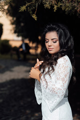 Naklejka premium Cute smiling woman with long wavy hair and makeup, attractive young girl enjoying summer in luxury white dress. Portrait of young woman standing on the park on sunny summer or spring day outside.