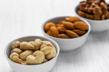 Cashews, almonds and walnuts in white glass bowls are located diagonally on a white wooden background, side view from above