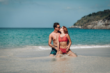Fototapeta premium Loving couple sitting on the wet sand near the ocean kissing and hugging; honeymoon concept.