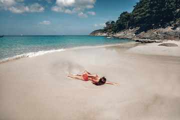 Fototapeta na wymiar Sexy girl in red bikini sunbathing on wet sand by the clear ocean, sunny day, rocks, trees; sunbathing concept.