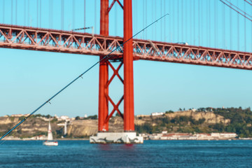 fishing pole against red bridge on the bank of Tagus river. Suspension Bridge over river on background.