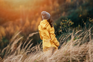 Young female with yellow raincoat in tall grass during sunset light - Windy outdoors weather