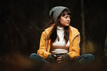 Female traveler in yellow rain coat sitting next to camp fire in forest with hot drink in hand / Dark autumn mood in the woods.