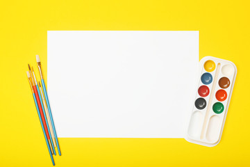 Blank paper with brushes and paints on yellow background. Artist workplace concept with copy space for your ideas. Flat layout, top view.