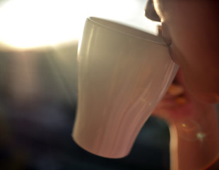 Woman drinks from a white cup at sunset background, close-up