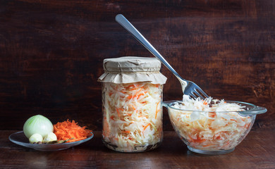 Homemade sauerkraut with carrots in a glass jar and bowl on a dark wooden background. Fermented food. Marinated vegetables