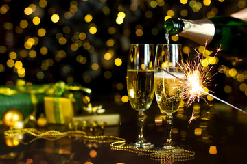 Glass is filling with champagne, sparkler is burning. New Year golden decor, balls, presents are on table. Festive decorative garland with yellow light bulbs are shining on background. Christmas mood.