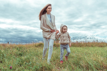 Happy family, mom woman with little boy 4-5 years old on weekend walk in park field, background green grass. Warm casual wear, hooded sweater. Caring support love and parenting hold hands.