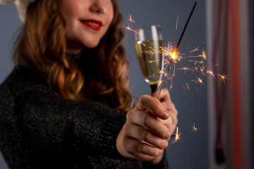 Attractive cheerful girl with curly hair in Christmas hat smiling, holding sparkler and drinking champagne, isolated on gray background