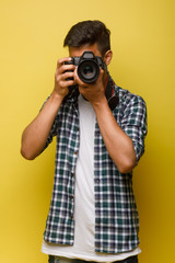 Handsome and confident indian man photographer with a large professional camera taking pictures photo shooting on the on the yellow background. Lifestyle.