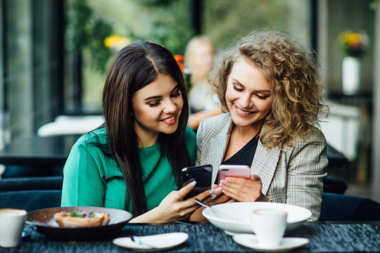 Two Girl Friends Spending Time Together Drinking Coffee In The Cafe, Having Breakfast And Dessert. Youth, Lifestyle, Communication Concept, Using Their Phones.