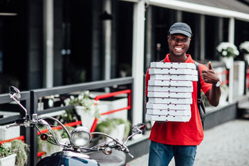 I recommend eat this delicious pizza! Cheerful busy man courier delivers appetizing fast food in cardboard container, keeps thumb up, shows like gesture, drives moped.