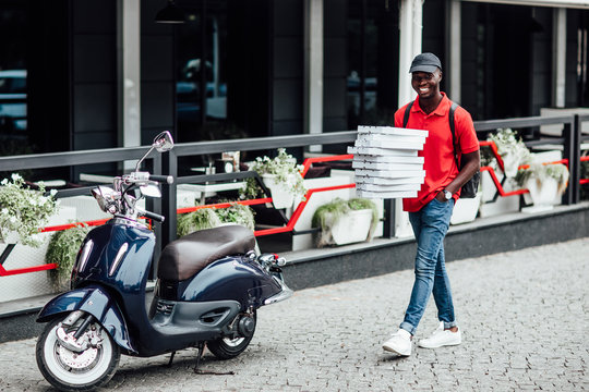 Busy deliveryman being in hurry, carries cardboard boxes with pizza, delivers to customers, poses on blue scooter, wears helmet, white t shirt and sportshoes, spreads legs, has happy expression.