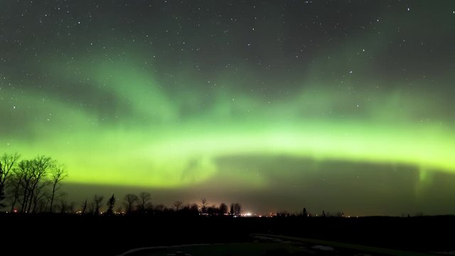 Time lapse video of Aurora (Northern Lights), stars, and clouds above a small town in the Canadian prairies.  A few clouds drift through the scene and reflect the light from the town.