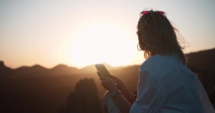 Pleased young woman enjoying bright sunset in beautiful mountain landscape. Modern girl tourist with smartphone on cliff top overlooking panoramic nature at sunset.