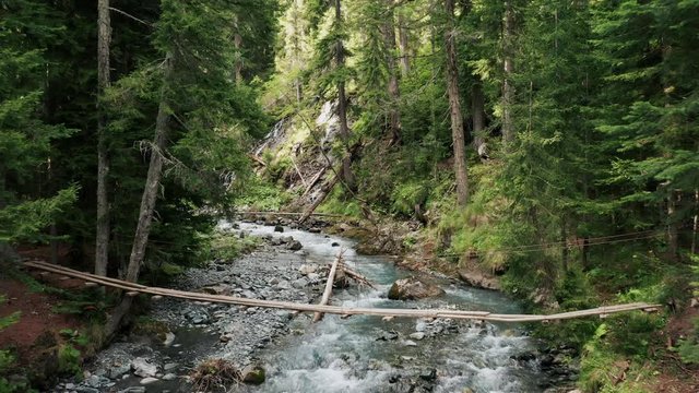 Picturesque suspension wooden bridge over stormy flow of crystal clear transparent water of mountain river; fabulous environment of wild natural places; eco-friendly construction in conservation area