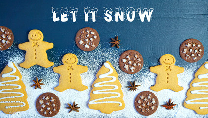 Christmas holiday flat lay composition of gingerbread and chocolate cookies on dark wood background table dusted powdered confectionery sugar, with copy space and Let It Snow text greeting.